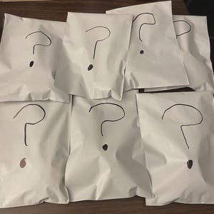 May include: Seven white paper packages, each with a large black question mark drawn on the front. The question marks are hand-drawn, and each package is sealed. The packages are arranged in a slightly overlapping pattern.