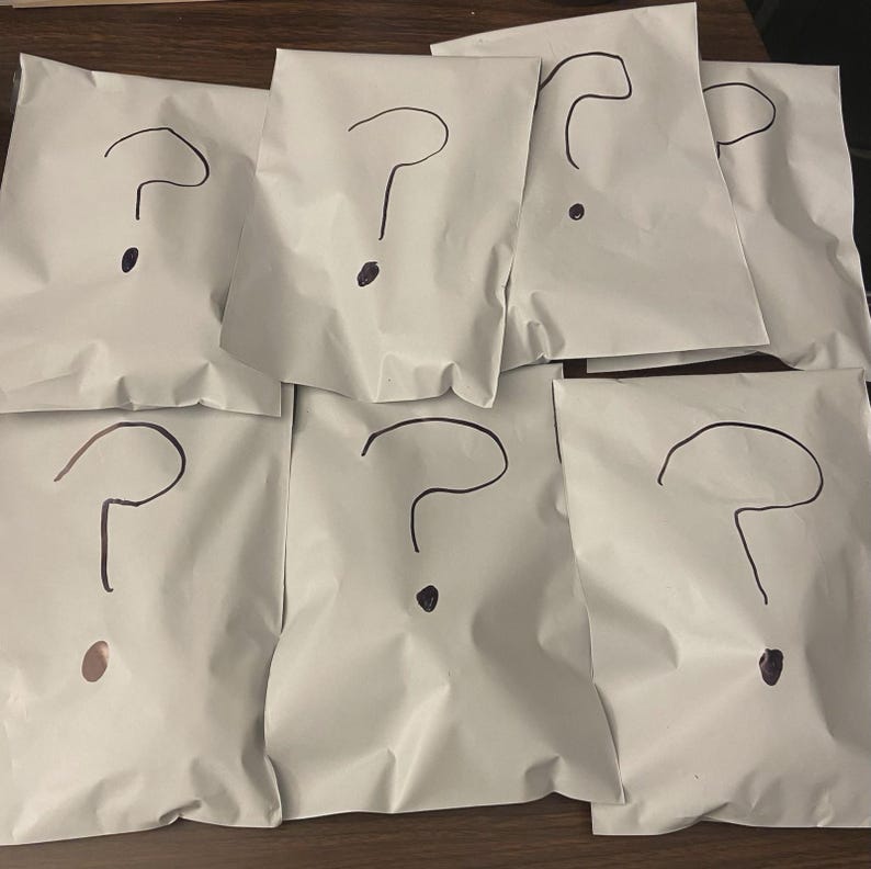 May include: Seven white paper packages, each with a large black question mark drawn on the front. The question marks are hand-drawn, and each package is sealed. The packages are arranged in a slightly overlapping pattern.