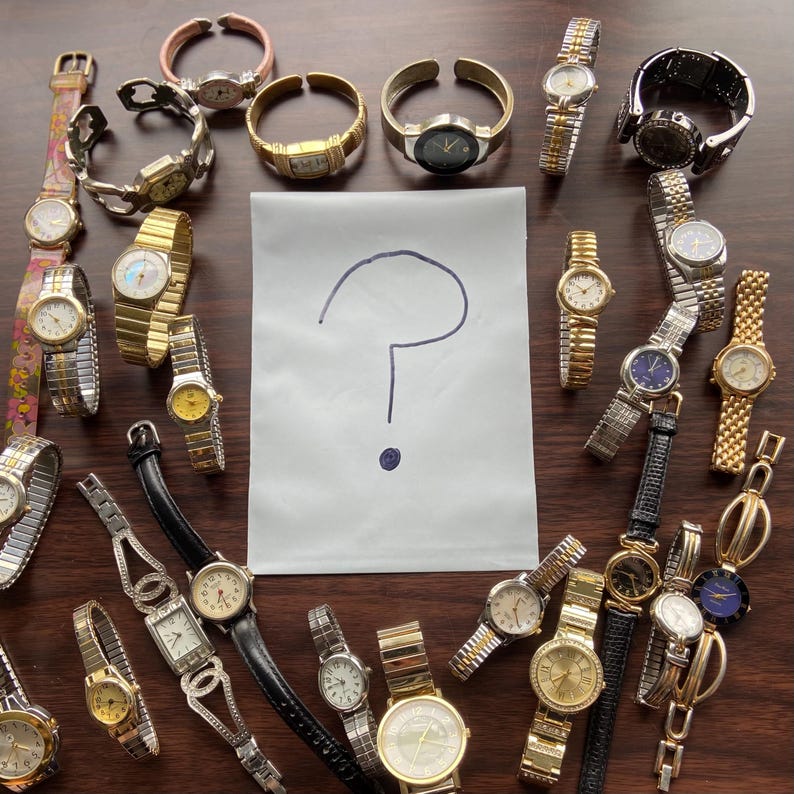 Op de afbeelding: Een verzameling diverse polshorloges met verschillende bandstijlen en wijzerplaatontwerpen. De horloges hebben gouden, zilveren en zwarte banden, sommige met decoratieve elementen. Een papier met een vraagteken staat in het midden.