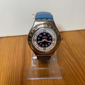 May include: A Swatch Swiss watch with a blue leather band. The watch face is black with white and red accents, featuring the Swatch Swiss logo. The watch is displayed on a clear plastic stand.