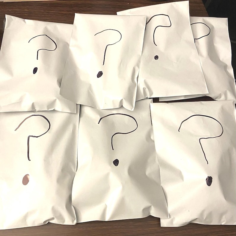 Op de afbeelding: Zeven witte papieren pakketten, elk met een zwart vraagteken. De pakketten liggen op een houten oppervlak. De vraagtekens zijn getekend met een dikke stift.