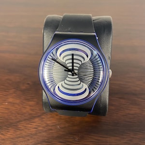 Puede incluir: Un reloj Swatch Swiss con correa negra y bisel azul. La esfera del reloj presenta un diseño en espiral plateado y blanco. El reloj se muestra sobre un soporte negro para relojes, sobre una superficie de madera marrón.