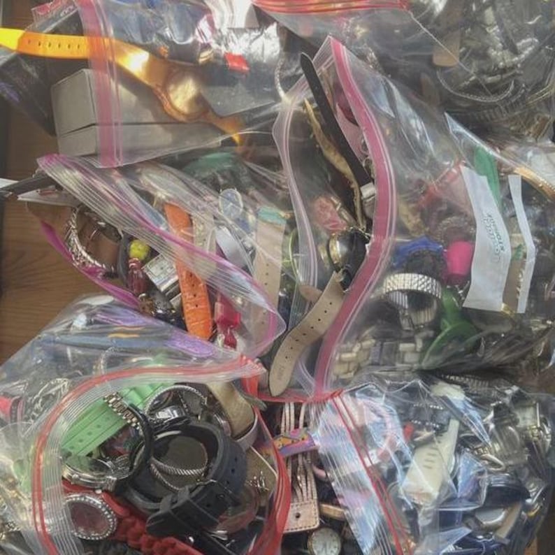 Op de afbeelding: Assortiment horloges in doorzichtige plastic zakken. De horloges hebben verschillende bandkleuren, waaronder zwart, bruin, oranje en groen. Sommige horloges hebben metalen banden, andere leren of plastic bandjes. De afbeelding toont een verscheidenheid aan horlogestijlen.