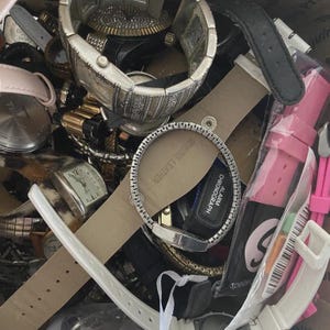 Op de afbeelding: Een verzameling verschillende polshorloges, met een verscheidenheid aan stijlen en kleuren. De horloges omvatten metalen banden, leren bandjes en digitale displays. Sommige horloges hebben zilver- en goudtinten, terwijl andere roze en zwarte bandjes hebben.