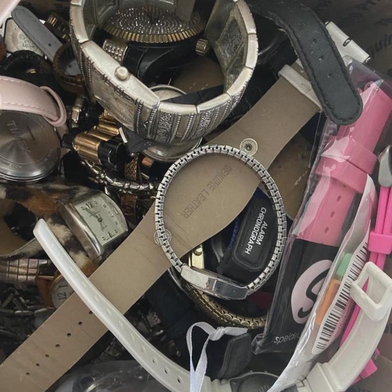 Op de afbeelding: Een verzameling verschillende polshorloges, met een verscheidenheid aan stijlen en kleuren. De horloges omvatten metalen banden, leren bandjes en digitale displays. Sommige horloges hebben zilver- en goudtinten, terwijl andere roze en zwarte bandjes hebben.