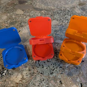 Könnte beinhalten: Drei runde, scharnierartige Kunststoffbehälter in Blau, Orange und Rot. Die Behälter sind geöffnet und geben den Innenraum frei. Die Deckel haben eine quadratische Form und sind mit einem Scharnier an der Basis befestigt. Die Behälter scheinen 3D-gedruckt zu sein.