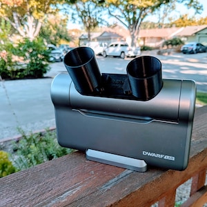 May include: A gray DwarfLab device with two black cylindrical attachments. The device is resting on a wooden railing outdoors, with the text "DwarfLab" visible. The background includes trees, houses, and parked cars.