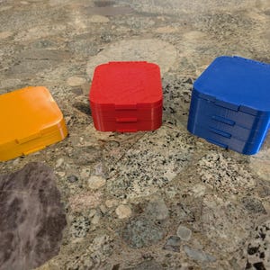 May include: Three small, square, plastic boxes in primary colors: orange, red, and blue. Each box has a hinged lid and a textured surface. The boxes are arranged on a speckled, gray surface.