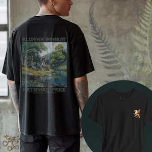 Könnte beinhalten: Schwarzes T-Shirt mit Landschaftsmotiv und dem Text "ELWYNN FOREST NATIONAL PARK EASTERN KINGDOMS". Ein kleineres schwarzes T-Shirt zeigt ein goldenes Greifen-Emblem. Die T-Shirts sind aus weichem Material.