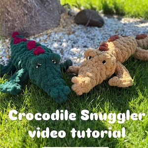 May include: Two crocheted crocodile plushies, one green and one brown, lying on a bed of green grass. The text "Crocodile Snuggler video tutorial" and "Level: Advanced" is displayed below the plushies.
