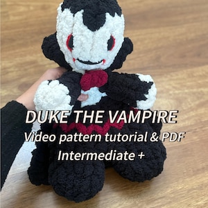 May include: A crochet vampire doll in black, white, and red. The doll features a black head and body, a white face, and red accents. The image includes the text "DUKE THE VAMPIRE" and "Video pattern tutorial & PDF Intermediate +".