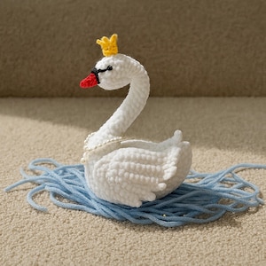 Puede incluir: Un cisne blanco tejido a mano con ganchillo, adornado con una corona amarilla, pico rojo y ojos negros. El cisne tiene un collar de perlas y descansa sobre hilo azul, un artículo decorativo encantador.