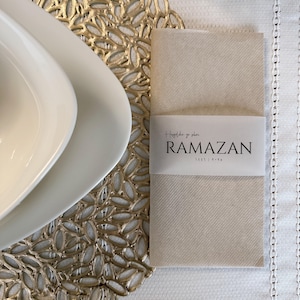 Op de afbeelding: Een beige papieren plaatskaart met de tekst "Hoşgeldiniz ya dahları" en "RAMAZAN 1443 | 2022" in zwart gedrukt. De plaatskaart ligt op een gouden placemat met een decoratief patroon.