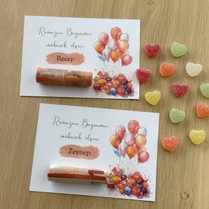 May include: Two white cards with a watercolor illustration of colorful balloons and candy. The cards have the text "Ramazan Bayramin mubarek olsun" and the names "Recep" and "Zeynep" written on them. Rolled up bills are placed on the cards.  There are also several heart-shaped candies in various colors scattered around the cards.