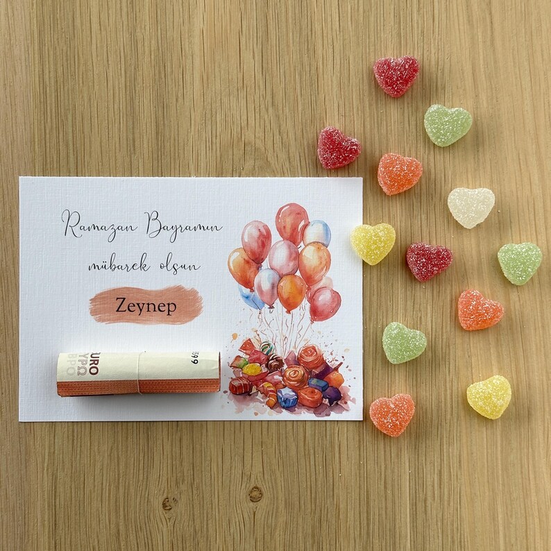May include: A white card with the text "Ramazan Bayramin mubarek olsun Zeynep" and a watercolor illustration of colorful balloons and candy. A rolled up bill of money is on the card. The card is surrounded by heart-shaped candies in various colors.