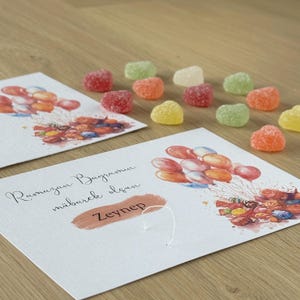 May include: A white card with a watercolor illustration of colorful balloons and candy. The card has the text "Ramazan Bayramınız m&uuml;barek olsun" and the name "Zeynep" written on it. The card is surrounded by heart-shaped candies in various colors.