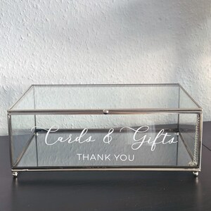 Può includere: Una scatola rettangolare di vetro per biglietti e regali con una cornice in metallo argentato. La scatola ha un coperchio incernierato ed è decorata con le parole "Cards & Gifts" e "Thank You" in scrittura bianca. La scatola poggia su quattro piedini.