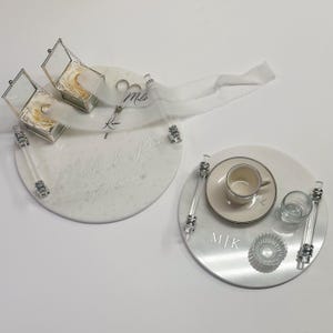 May include: Two white marble-effect trays with clear handles and silver accents. One tray holds two ring boxes, the other a teacup, glass, and decorative item. The trays are personalized with names and a date.