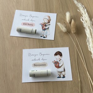 May include: Two white cards with illustrations of children holding baskets of flowers. The cards have the text "Ramazan Bayraminiz mubarek olsun" and the names "Elif Serra" and "Bunyamin". Rolled up bills of money are placed on each card.