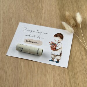 May include: A white card with the text "Ramazan Bayramin mubarek olsun" and the name "Bunyamin" written in cursive. A cartoon illustration of a boy holding a basket of flowers is on the right side of the card. A rolled-up euro bill is on the left side of the card.