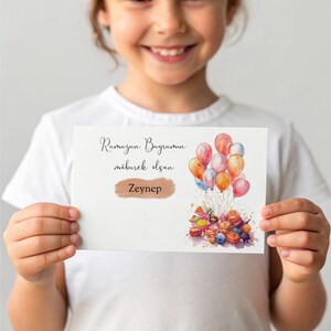 May include: A white card with the text "Ramazan Bayramin" and "mubarek olsun" in brown lettering. The card also has a gold name tag with the name "Zeynep" on it. The card has a watercolor illustration of colorful balloons and candy.