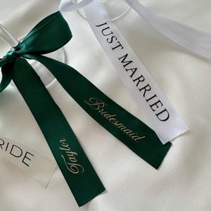 Può includere: Primo piano di nastri nuziali. Un nastro verde scuro è legato a fiocco, con una lunga coda che recita "Taylor" in oro. È visibile anche un nastro bianco con "Just Married" in testo nero. I nastri sono su sfondo bianco.