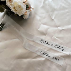 Könnte beinhalten: Ein weißes Band mit ausgefransten Kanten, auf dem die Namen "Meltem & Kubilay" und das Datum "12 oktober 2025" in schwarzer Schrift stehen. Das Band liegt neben einem Strauß weißer und cremefarbener Rosen.