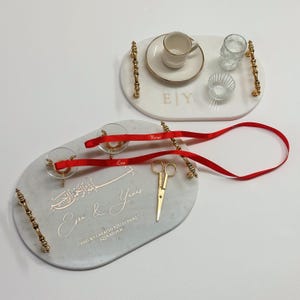 May include: Two marble serving trays with gold handles and accents. One tray holds a teacup, saucer, and small glass items. The other tray has a pair of glasses with red ribbons and gold scissors. Text is visible.