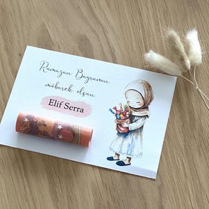 May include: A white card with the text "Ramazan Bayraminiz mubarek olsun" and "Elif Serra" written on it. A cartoon illustration of a girl wearing a headscarf and holding a bag of candy is on the card. A rolled up bill of money is on the card.
