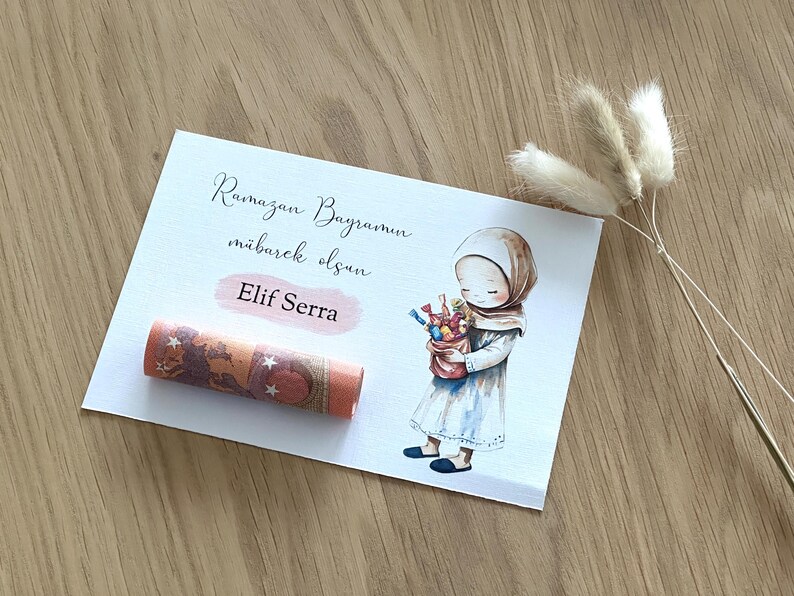 May include: A white card with the text "Ramazan Bayraminiz mubarek olsun" and "Elif Serra" written on it. A cartoon illustration of a girl wearing a headscarf and holding a bag of candy is on the card. A rolled up bill of money is on the card.