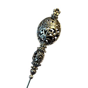 May include: A silver-toned hat pin with an ornate, filigree design. The pin features a decorative oval top with intricate cut-out patterns and a floral-like finial. The stem is slender and pointed, suitable for securing headwear.