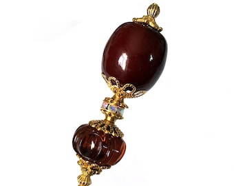May include: A gold and brown glass hat pin with a large, dark red glass bead at the top. The pin has a decorative, ornate design.