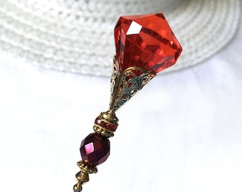 May include: A red glass pin with a gold filigree design and a dark red glass bead. The pin is on a white background.