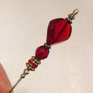 May include: A red glass bead pin with gold accents. The pin has a teardrop-shaped bead and a round bead.