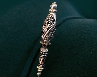 May include: A silver filigree pin with a decorative, ornate top and a long, thin needle. The pin is on a dark green background.
