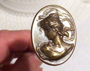 May include: An oval-shaped metal wax seal stamp featuring a raised bronze-toned relief of a profile portrait of a person with flowing hair, set against a smooth, iridescent, off-white mother-of-pearl background with a thin gold-toned metal border.