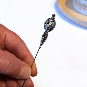 May include: An ornate silver-toned hat pin with a decorative filigree design. The pin is held in a hand against a white background, with a portion of a hat visible in the background.