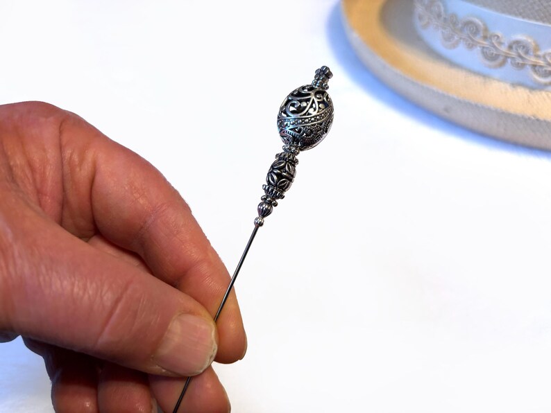 May include: An ornate silver-toned hat pin with a decorative filigree design. The pin is held in a hand against a white background, with a portion of a hat visible in the background.