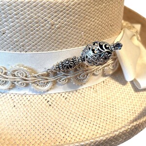 May include: A straw hat with a white ribbon and decorative silver embellishment. The hat features a wide brim and a decorative silver pin with an ornate design. The ribbon is adorned with a delicate, embroidered trim.