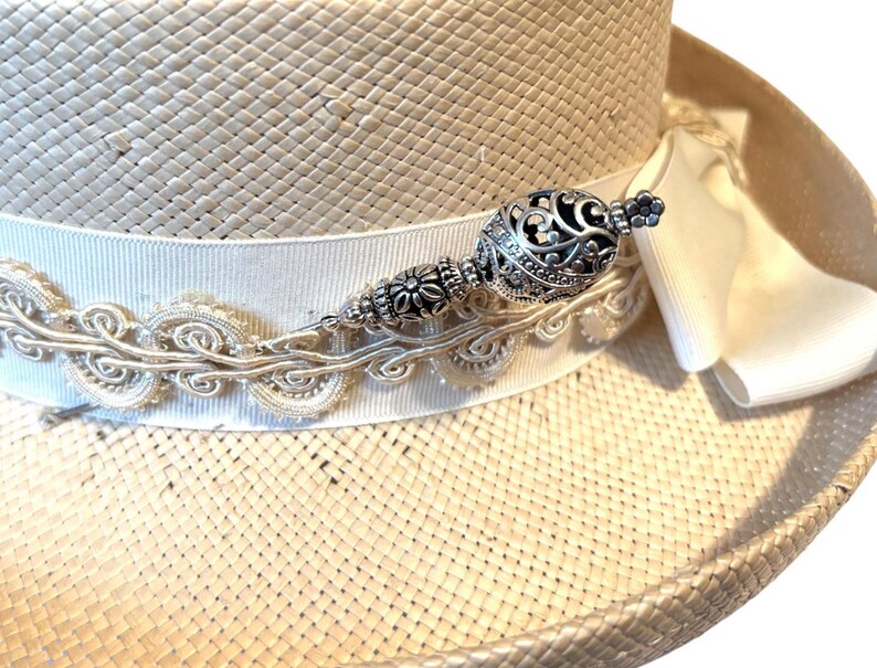 May include: A straw hat with a white ribbon and decorative silver embellishment. The hat features a wide brim and a decorative silver pin with an ornate design. The ribbon is adorned with a delicate, embroidered trim.
