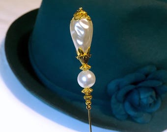 May include: A decorative hat pin with a pearlescent, teardrop-shaped top and a round pearl accent. The pin features gold-colored metal detailing and is set against a teal hat with a matching flower.