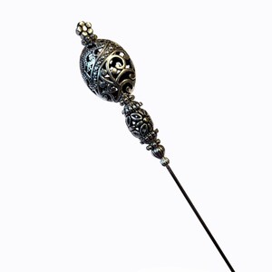 May include: A silver-toned hat pin with an ornate, filigree design. The pin features a decorative top with an oval shape and a smaller, floral-shaped top. The long, slender pin is ideal for securing hats or adding a touch of elegance.