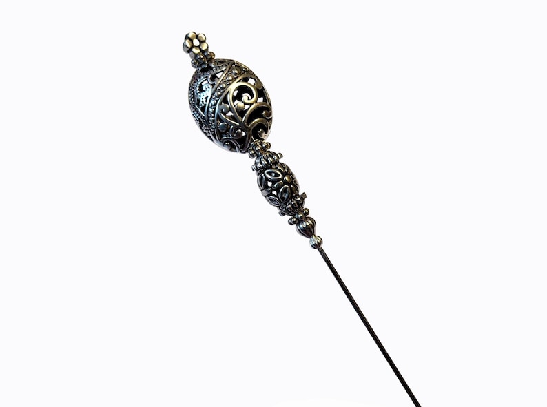 May include: A silver-toned hat pin with an ornate, filigree design. The pin features a decorative top with an oval shape and a smaller, floral-shaped top. The long, slender pin is ideal for securing hats or adding a touch of elegance.
