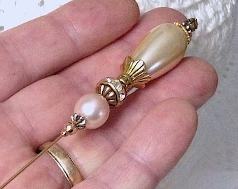 May include: A vintage hat pin with a pearlescent, teardrop-shaped top and a large, round pearl bead. The pin features gold-toned metal accents and small, sparkling rhinestones. The pin is held in a hand.