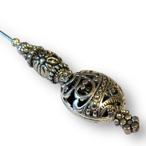 May include: Silver-toned decorative pin with an ornate, filigree design. The pin features a long, slender needle and a detailed, oval-shaped head with intricate patterns and a small cluster of beads at the end. The pin is likely used for decorative purposes.