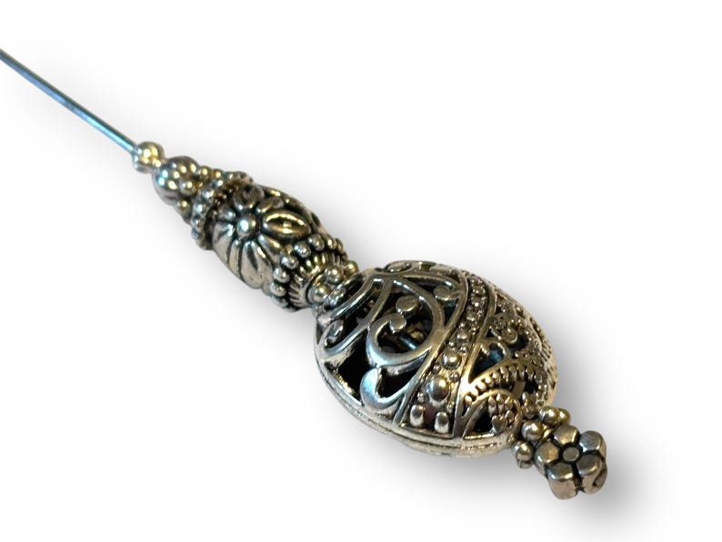 May include: Silver-toned decorative pin with an ornate, filigree design. The pin features a long, slender needle and a detailed, oval-shaped head with intricate patterns and a small cluster of beads at the end. The pin is likely used for decorative purposes.