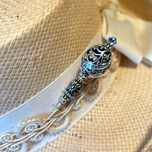 May include: A straw hat with a cream-colored ribbon and a decorative silver hat pin. The hat pin features an ornate design with a filigree ball and floral accents. The hat is a light beige color.