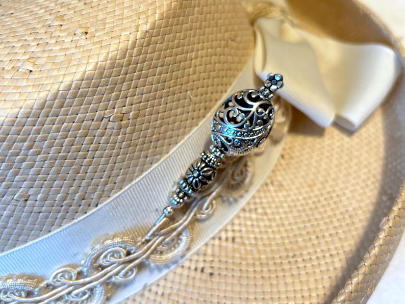 May include: A straw hat with a cream-colored ribbon and a decorative silver hat pin. The hat pin features an ornate design with a filigree ball and floral accents. The hat is a light beige color.