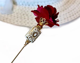 May include: A decorative hat pin featuring a deep red fabric rose with a gold-colored crown and accents. The pin has a white and gold-colored decorative element and a long, slender gold-colored pin.
