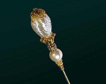 May include: A gold and white pearl pin with a decorative design. The pin has a large, pearlescent, round bead at the top and a smaller, round bead below it. The pin is attached to a gold metal stem.